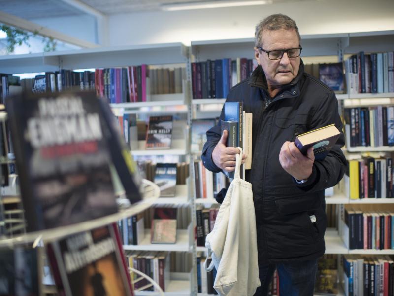 En mand læser bagsideteksten på en bog, mens han har to andre bøger under armen. Han står foran en bogreol.